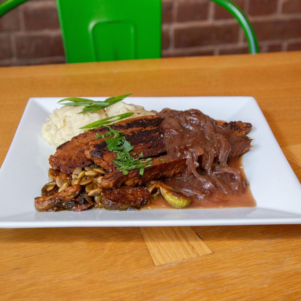 Grilled meat topped with caramelized onions, mashed potatoes, roasted vegetables, pumpkin seeds, and fresh herbs on a white plate.