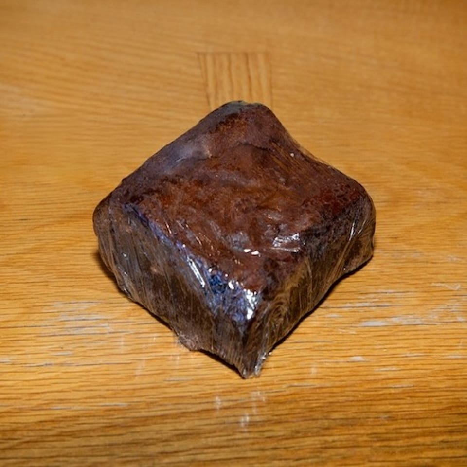 Glossy, dark-brown square-shaped food item with an irregular peaked top resting on a wooden table.