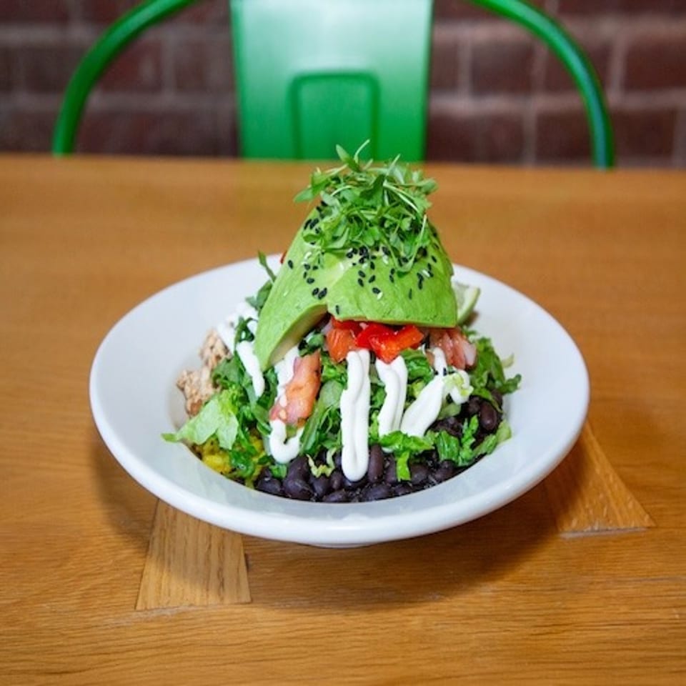 Layered vegetarian bowl with black beans, greens, vegetables, creamy dressing, avocado, and fresh sprouts on a white plate.