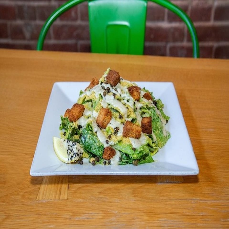 Caesar salad topped with croutons, creamy dressing, black seeds, and lemon wedge on a square white plate.