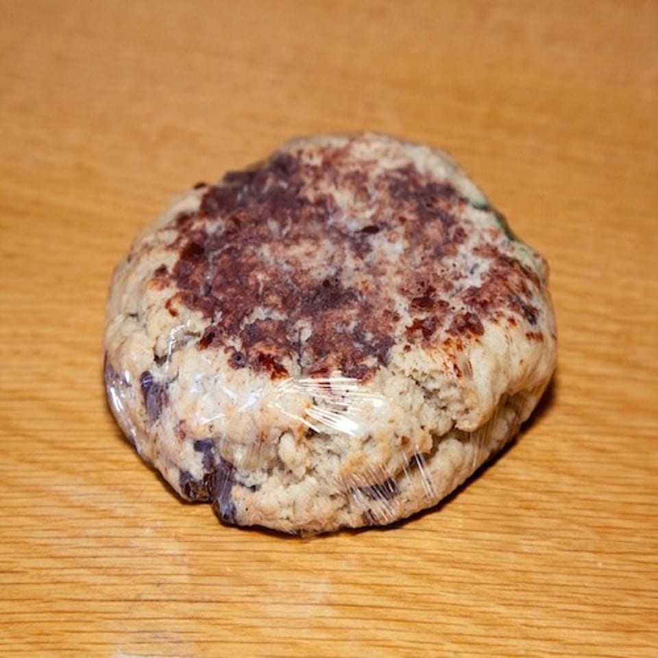 Round, rustic baked pastry with dark berry filling, wrapped in plastic on a wooden tabletop.