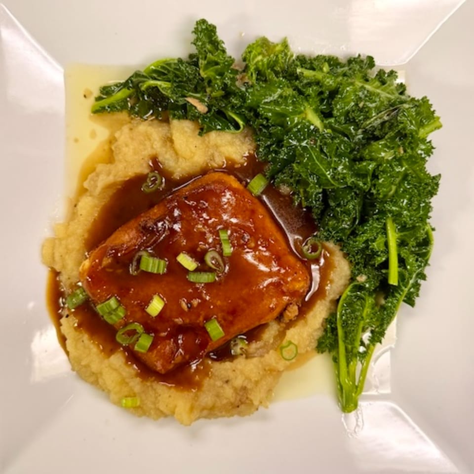 Glazed salmon topped with scallions, served over mashed potatoes with crispy kale on a white plate.