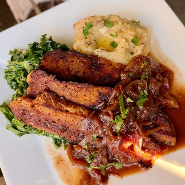 Vegan seitan slices with caramelized onions, mashed potatoes, and sautéed kale on a white plate.