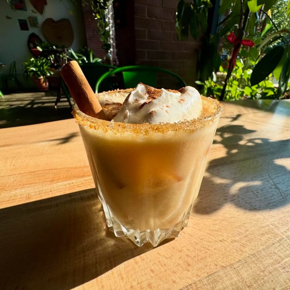 Vegan iced drink with whipped cream, cinnamon stick, and sugar rim on a sunlit wooden table.