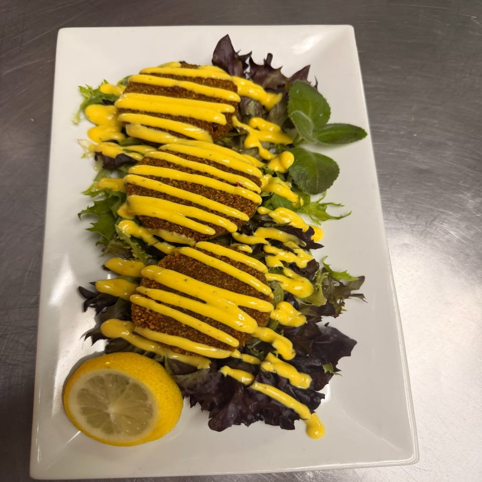 Crispy breaded fillets drizzled with yellow sauce over mixed greens, garnished with lemon on a white rectangular plate.