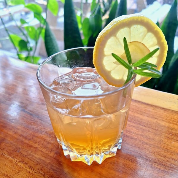A refreshing vegan cocktail with ice, a lemon slice, and rosemary garnish on a wooden table.